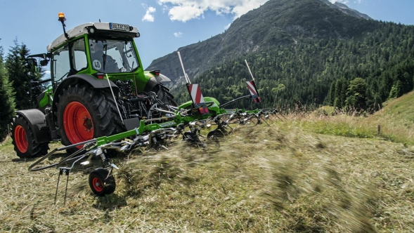 Fendt Twister hay tedder The green Fendt Twister with a green Fendt tractor and red rims tedding hay on a mountain meadow with a blue sky and mountain in the background.