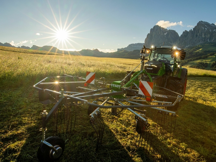 Fendt Former The green Fendt Former Alpine rake stands in front of a green Fendt tractor in a mountain meadow. In the background, the sun is slowly setting, the sky is blue and the mountains can also be seen