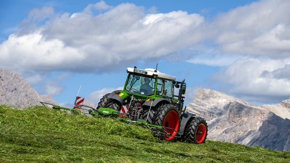 Fendt Alpine tedder The Fendt Alpine tedder and the green Fendt tractor with red rims in action on a mountain meadow with a blue sky, white clouds and mountains in the background.