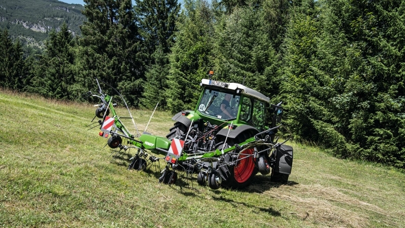 Fendt Twister hay tedder The folded Fendt Twister hay wagon is attached to a green Fendt tractor with red rims and driving across a meadow with a forest in the background