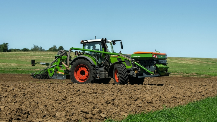 Der grüne Fendt 600 Vario mit roten Felgen und einer Drillmaschine als Anbaugerät fährt auf einem Acker. Im Hintergrund ist eine grüne Wiese und ein blauer Himmel zu sehen.