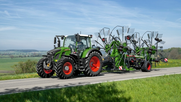 Fendt 600 Vario und Fendt Former Der grüne Traktor Fendt 600 Vario mit roten Felgen fährt mit dem grünen Fendt Former auf einer grauen Straße. Im Hintergrund ist eine große grüne Wiese und ein blauer Himmel.