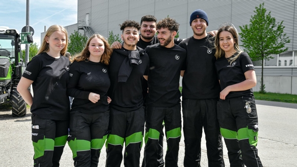 Seven Fendt employees, three female and four male, wearing the classic black Fendt work clothes with green elements, stand outside in front of a Fendt production building. A Fendt tractor can be seen in the background on the left.