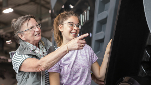 Two people are standing in front of a large machine. The older person points to a detail while the younger person looks attentively.