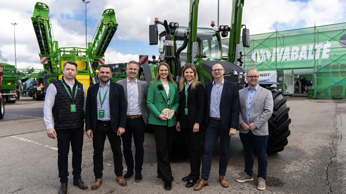 7 people, women in the centre, a woman with a green jacket holds an award in her hand, in front of a green Fendt tractor