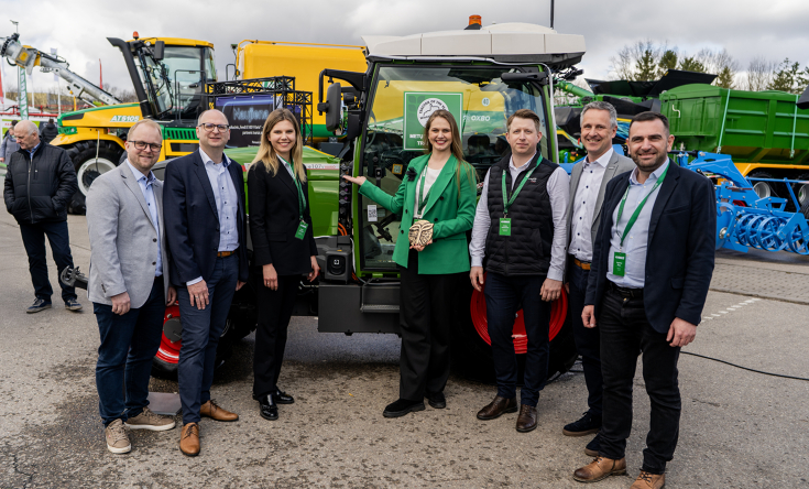 Ivabaltė exhibits Fendt e100 V Vario at the Ką pasėsi in the Baltic States 7 people, women in the centre, a woman with a green jacket holds a prize in her hand, in front of a green Fendt tractor