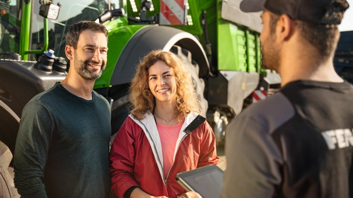 Two smiling people talking to a Fendt employee holding a tablet. A green tractor is clearly visible in the background. The scene takes place outdoors in the sunshine.