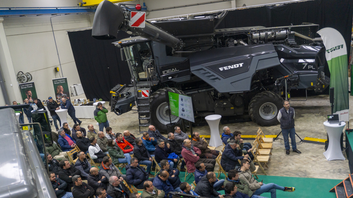 A room with many people sitting on chairs and looking towards the front, with a large Fendt IDEAL combine harvester with the side panel open on the side
