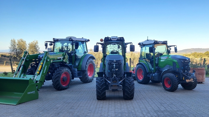 Three green Fendt 100 Vario tractors stand on the asphalt in the background, surrounded by grapevines.