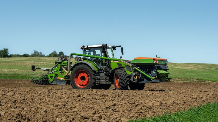 The green Fendt 600 Vario with red rims and a seed drill as an attachment driving in a field. A green meadow and a blue sky can be seen in the background.