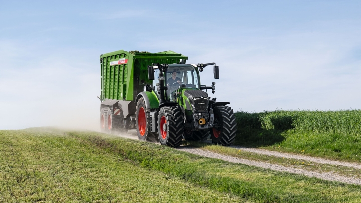 The green Fendt 600 Vario tractor with red rims and the green Fendt Tigo loader wagon driving along a small field road with a green meadow in the foreground and background.