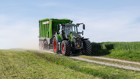 The green Fendt 600 Vario tractor with red rims and the green Fendt Tigo loader wagon driving along a small field road with a green meadow in the foreground and background.
