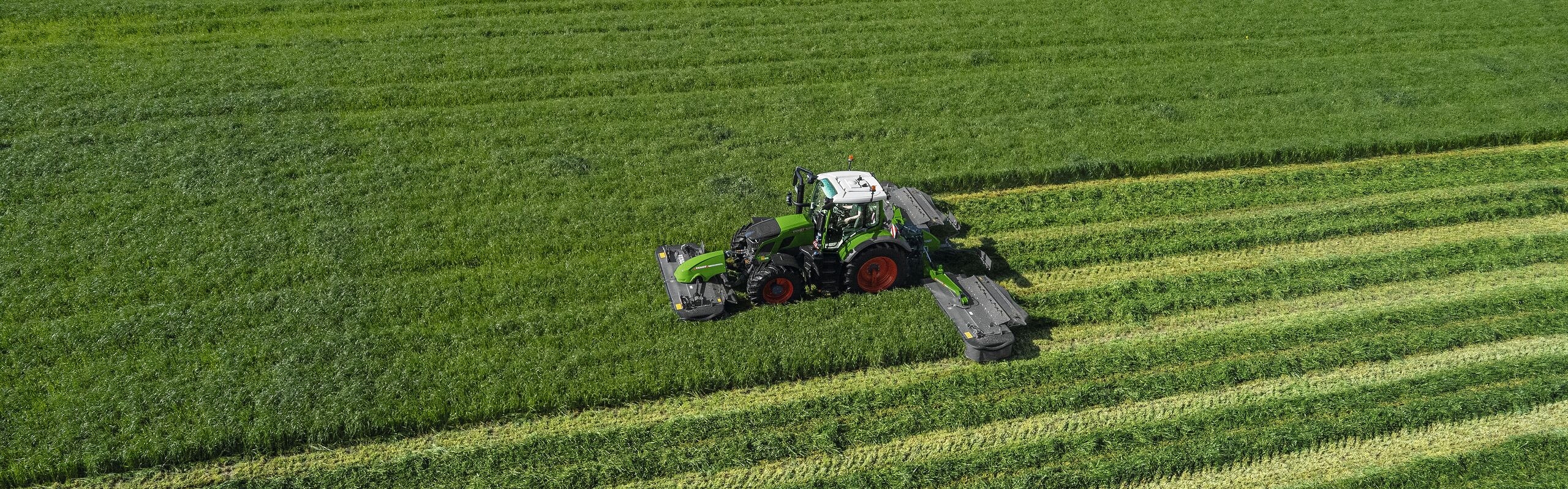 Bird's eye view of the green Fendt 600 Vario tractor in operation with the green Fendt Slicer mower on a large, green meadow.