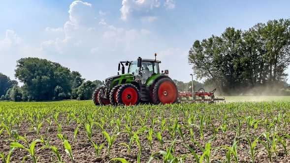 The green Fendt 600 Vario tractor with twin tyres in use on a cultivated field. Dark green trees and a light blue sky with white clouds can be seen in the background.