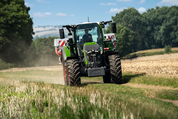 The green Fendt 600 Vario tractor with red rims travelling along a small country road with a green meadow in the foreground and a dark green forest and light blue sky in the background.