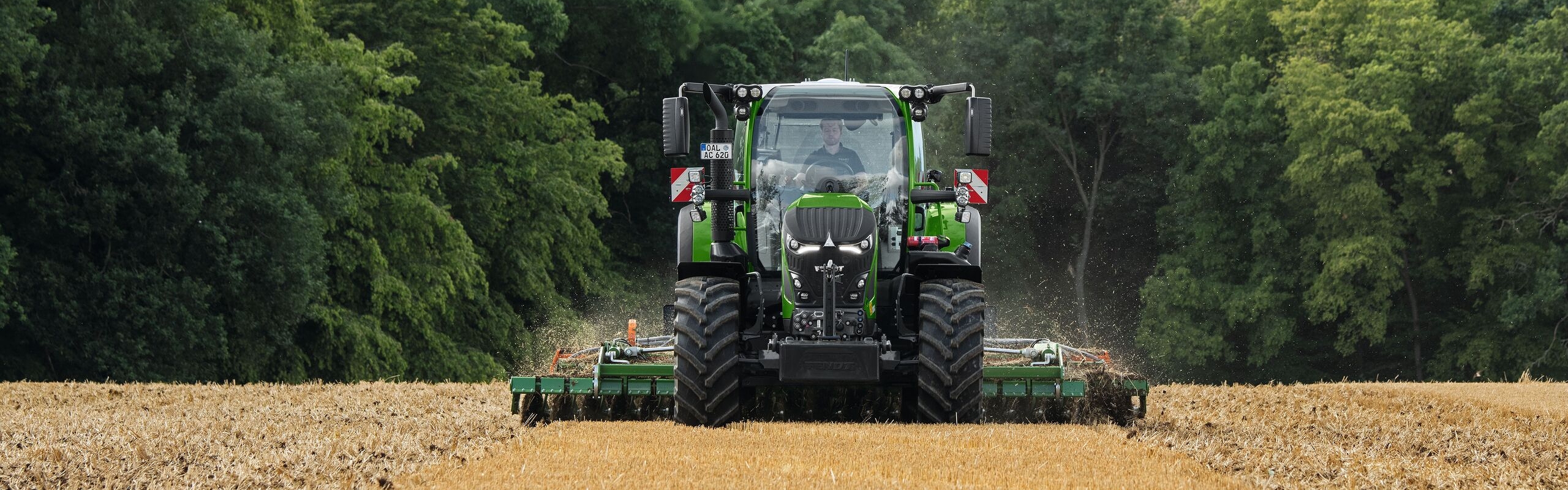 The green Fendt 600 Vario is in use with an implement in a brown field. In the background is a dark green forest and a cloudy sky.
