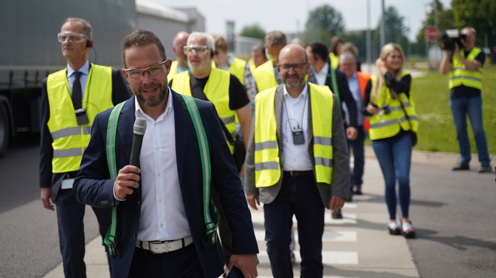 People are walking across a crosswalk on the factory premises and listening to Alexander Schenk speaking into a microphone.