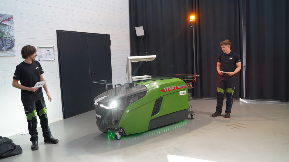 Two men (apprentices) stand next to the green Fendt dispensing station, one on the left and one on the right. The front lights, underbody lighting and rotating beacon are illuminated.