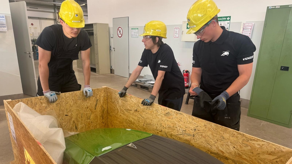 3 men (apprentices) are standing around an open box containing the Fendt bonnet, examining it and talking.