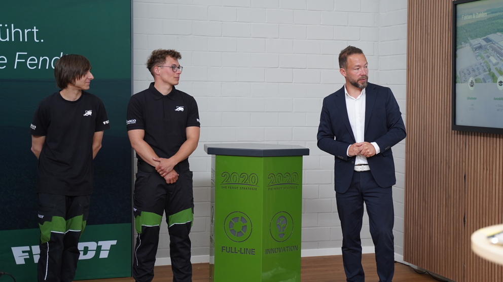 Apprentices and another man are standing in front of a wall. The two apprentices are looking at the man in the suit while he is speaking