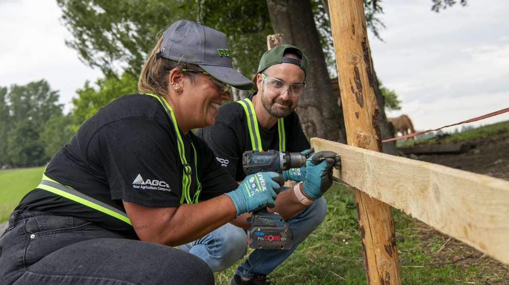 Fendt employees building a fence 2 people in protective work clothing, one of them a woman (front), squatting in front of a wooden fence. The woman is drilling into the fence while the man next to her watches in the background.