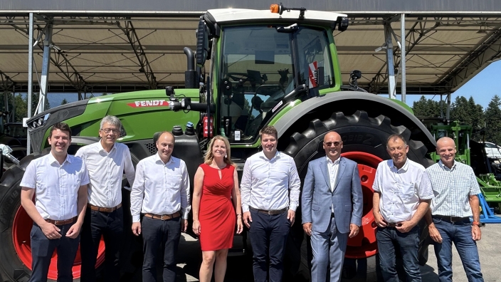 Group photo of 8 people, including 7 men and 1 woman in the middlewith a red dress, in front of a green Fendt tractor.