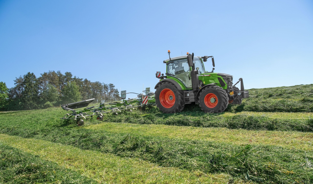 A Fendt 300 Vario with Fendt Twister tedder A green Fendt 300 Vario driving with a Fendt Twister tedder in grassland over hilly terrain.
