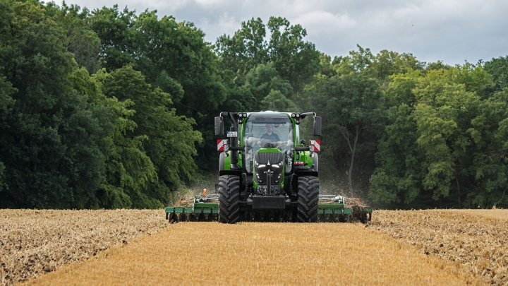 The green Fendt 600 Vario is in use with an attachment on a brown field. In the background is a dark green forest and a cloudy sky.