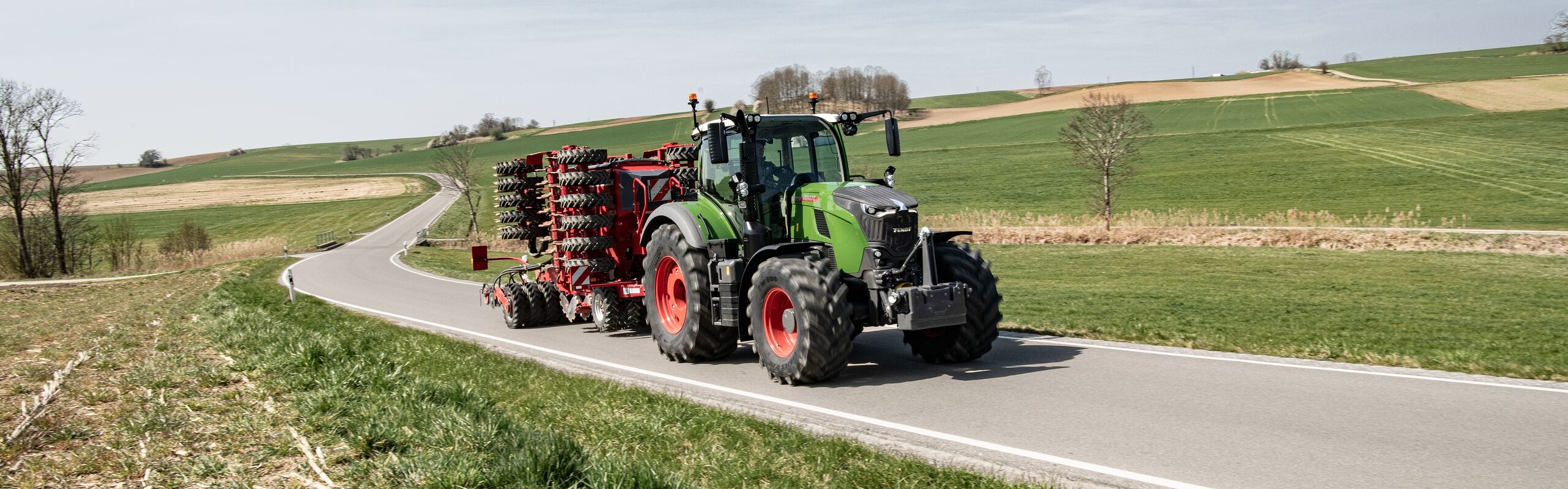 Een Fendt 700 Vario Gen7.1 met aangekoppelde aanbouwwerktuig op een landweg door een heuvelachtig voorjaarslandschap.