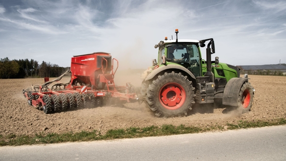 Een Fendt 700 Vario Gen7.1 met rode zaaimachine werkt of een stoffige akker tijdens zonnig weer.