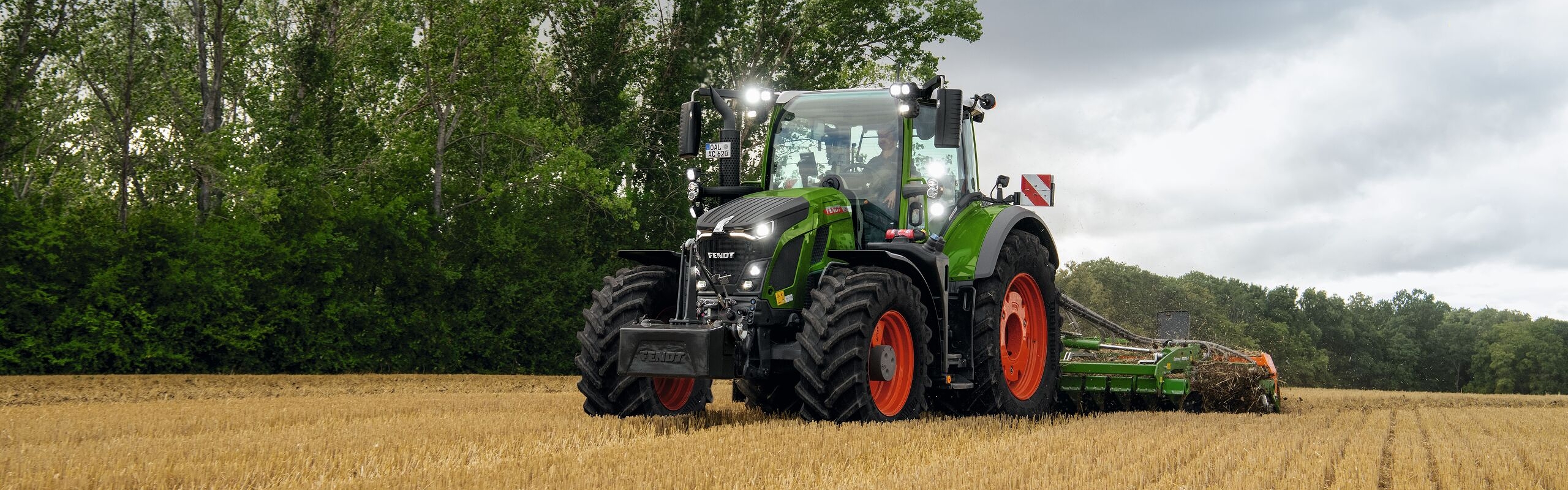 The green Fendt 600 Vario tractor with red rims in use on a field with trees in the background and a cloudy sky.
