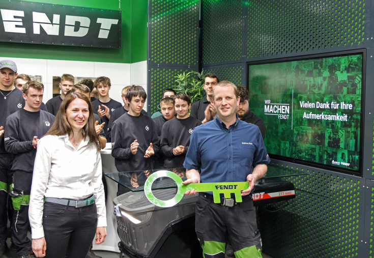 A woman on the left and a man on the right in the foreground, the man is holding a large green key with the inscription Fendt.