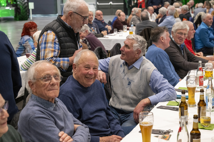 A group of older men are sitting at tables at the Fendt alumni meeting, some are chatting and smiling, drinks are in front of them.