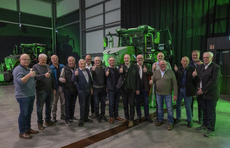 A group of men stand together and give a thumbs up, with tractors visible in the background.