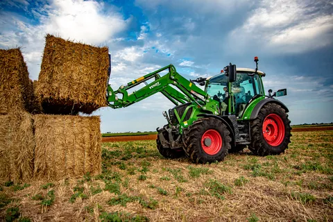 Un Fendt 300 Vario verde mentre impila balle di fieno su un prato verde con un campo marrone sullo sfondo.