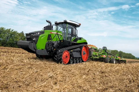 Vista laterale di un Fendt 1100 MT Vario con un attrezzo verde in un campo di stoppie. Sullo sfondo si vedono un bosco verde e un cielo azzurro con alcune nuvole sparse.