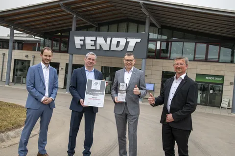 Foto di gruppo di Claus Meyer, Carsten Matthäus, Walter Wagner e Helmut Süß davanti al Fendt Forum. Tengono in mano il premio Fendt 1100 Vario MT e alzano il pollice in segno di approvazione.