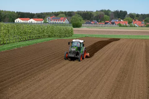 Un Fendt e107 Vario lavora con una fresa in un campo vicino a un frutteto; sullo sfondo si vedono le case con gli impianti fotovoltaici sui tetti.