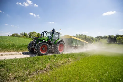 Un Fendt 500 Vario verde traina due rimorchi verdi con teloni gialli su una strada sterrata, sollevando una nuvola di polvere.