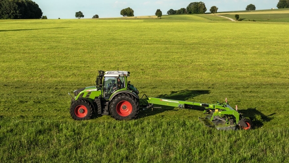 Fendt Slicer trainata in una prateria Fendt Slicer trainata con carrello di trasporto su un grande prato durante la falciatura. Sullo sfondo si vedono alberi e arbusti isolati.