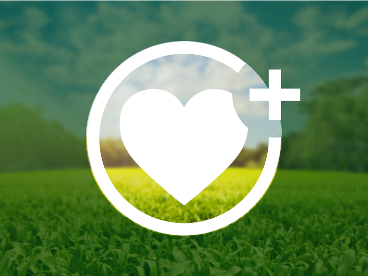 Un cuore bianco si trova all’interno di un cerchio, con un “più” sul lato destro. Lo sfondo mostra un prato verde sotto il cielo blu, che al di fuori del cerchio è oscurato da uno strato verde scuro, in parte trasparente.