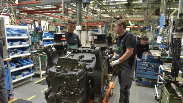 Diversi lavoratori in un capannone industriale montano delle grandi macchine con ruote dentate e componenti visibili. Indossano indumenti da lavoro e guanti. Sullo sfondo si vedono delle mensole con dei contenitori blu, oltre ad altre postazioni di lavoro