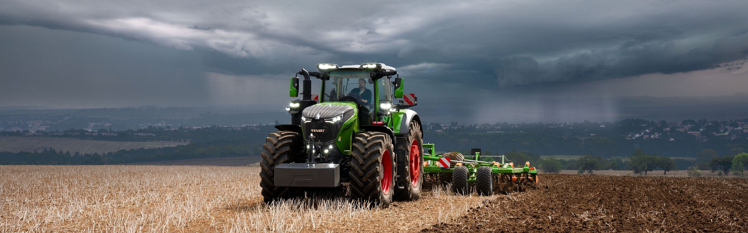 Davanti a nuvole temporalesche che si addensano all'orizzonte, il trattore Fendt 1000 Vario continua a svolgere con efficacia il suo lavoro nei campi.