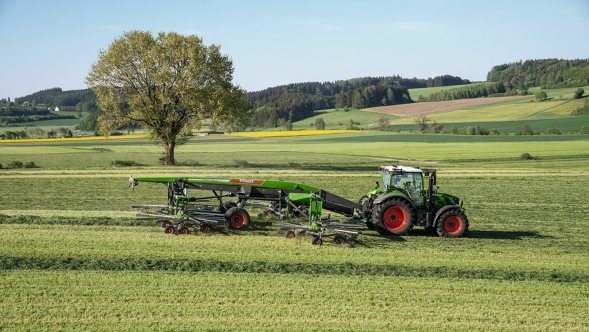 Un trattore Fendt verde con cerchi rossi e un Fendt Former 14055 PRO agganciato durante la ranghinatura su un prato verde estivo
