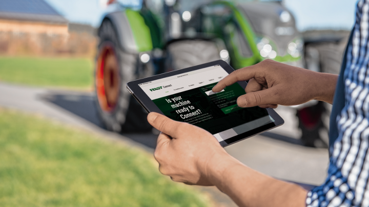 Vista ravvicinata di un tablet in mano a un agricoltore che controlla i dati centrali della propria macchina tramite Fendt Connect. Sullo sfondo si vede un trattore verde a marchio Fendt leggermente sfumato.