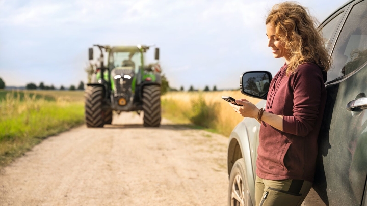 Una donna è appoggiata a un'auto e guarda il suo cellulare, sullo sfondo si vede un trattore Fendt.