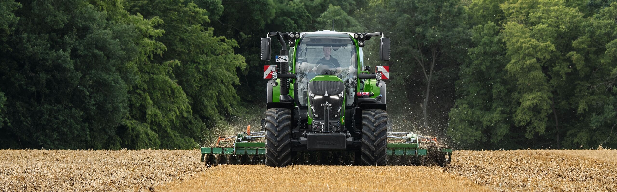 Il Fendt 600 Vario verde durante l’utilizzo con un attrezzo su un campo marrone. Sullo sfondo una foresta verde scuro e un cielo coperto di nuvole.