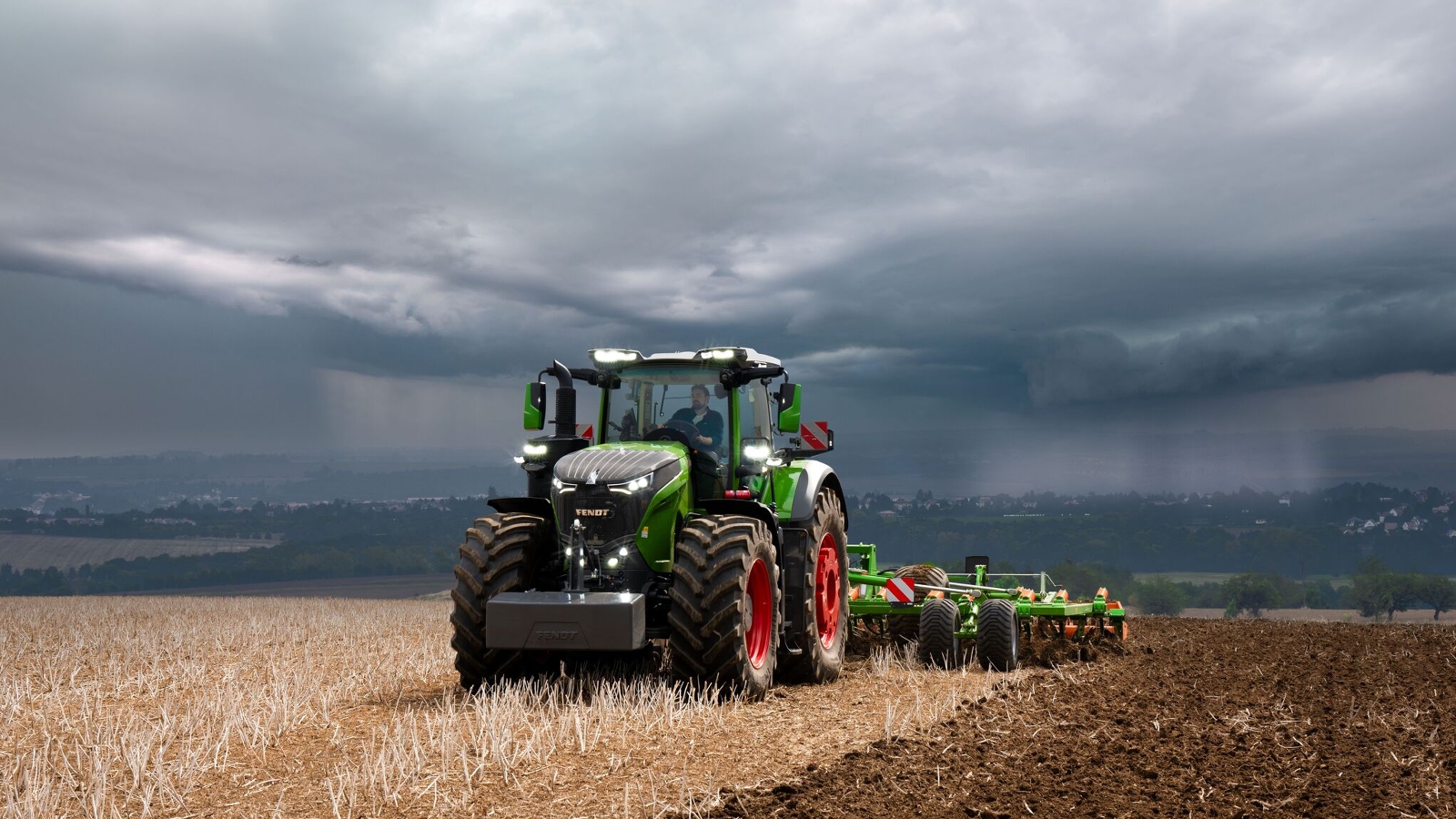 Davanti a nuvole temporalesche che si addensano all'orizzonte, il trattore Fendt 1000 Vario continua a svolgere con efficacia il suo lavoro nei campi.