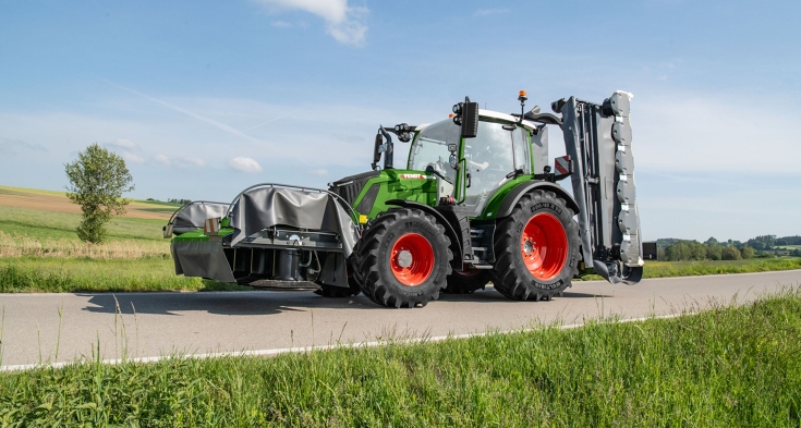 un Fendt 300 Vario verde procede con una falciatrice anteriore e posteriore Fendt Slicer su una strada.