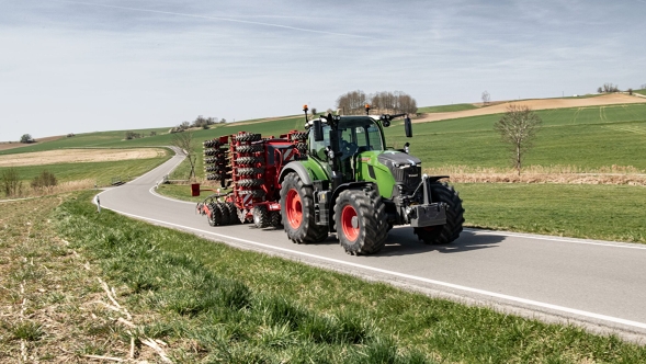 Il Fendt 700 Vario Gen7.1 con attrezzo agganciato procede su una strada di campagna attraverso un paesaggio collinare primaverile.